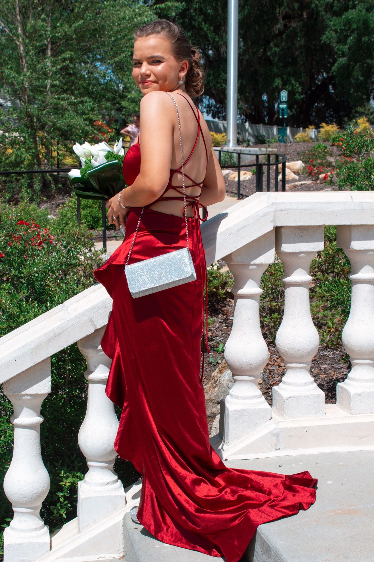 Young woman in backless red velvet prom gown poses on white balustrade holding white flower bouquet.