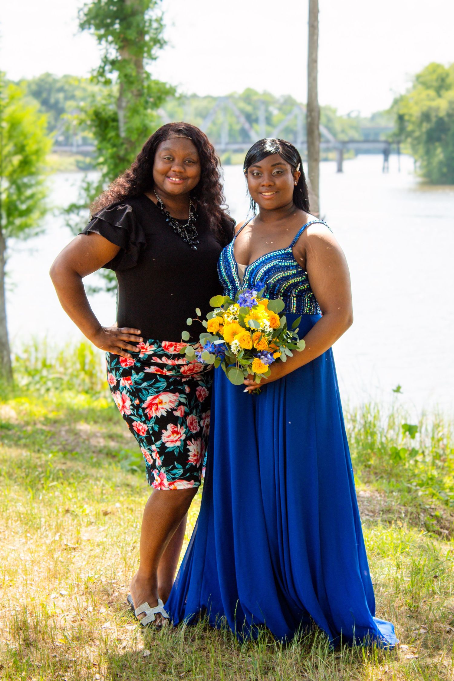 Two young women pose outdoors near a river, one wearing a stunning blue prom dress holding a yellow bouquet.