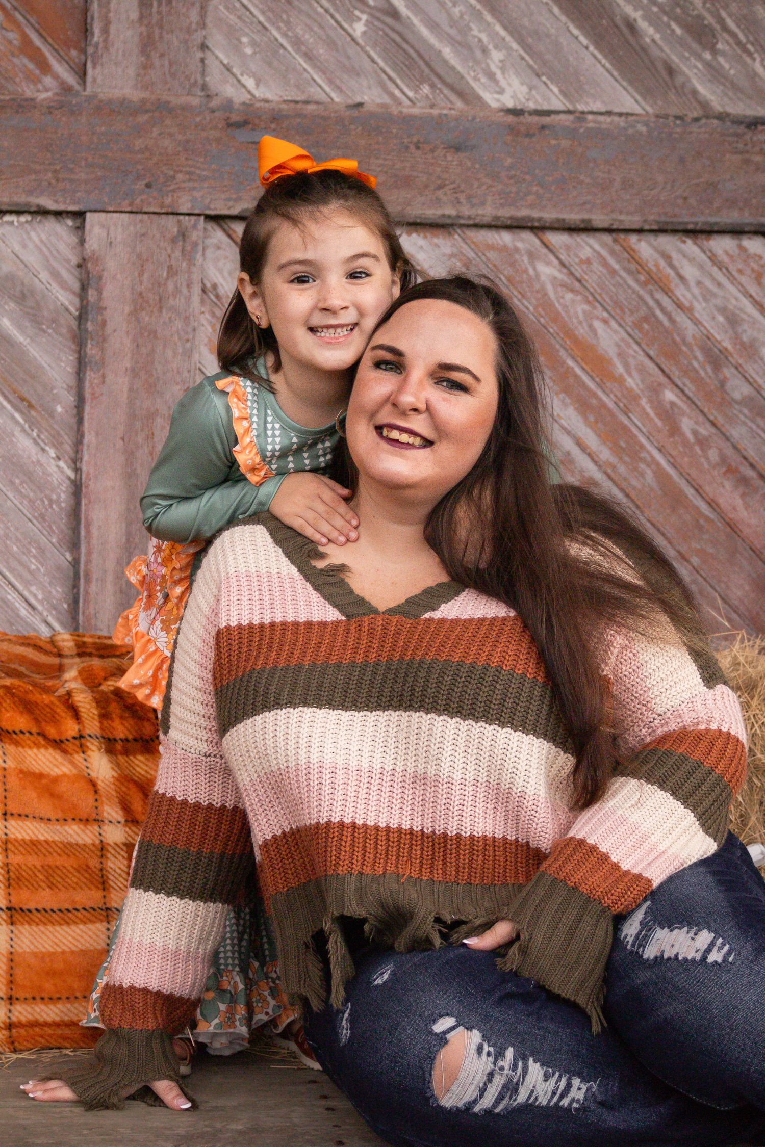 Young girl in fall outfit hugs smiling mother in striped sweater against rustic wooden barn backdrop.