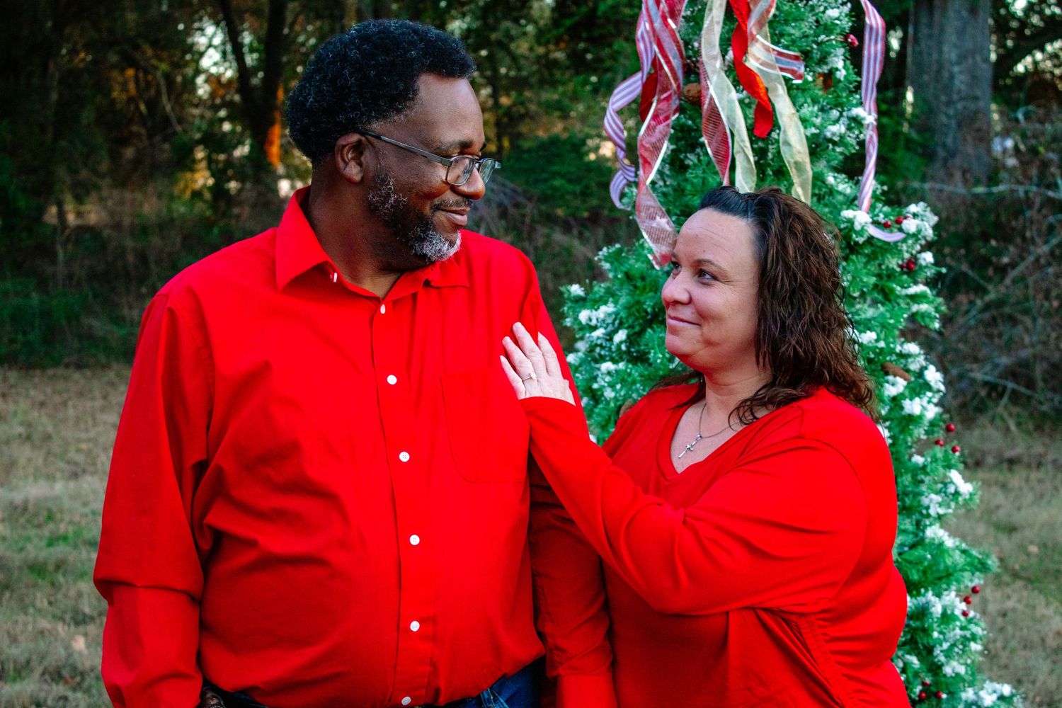 Couple in matching red shirts smiling at each other near a decorated Christmas tree outdoors.