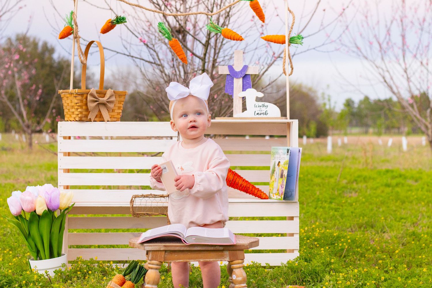 Adorable baby girl in white bow sits on stool at Easter photo backdrop with carrots and tulips outdoors.
