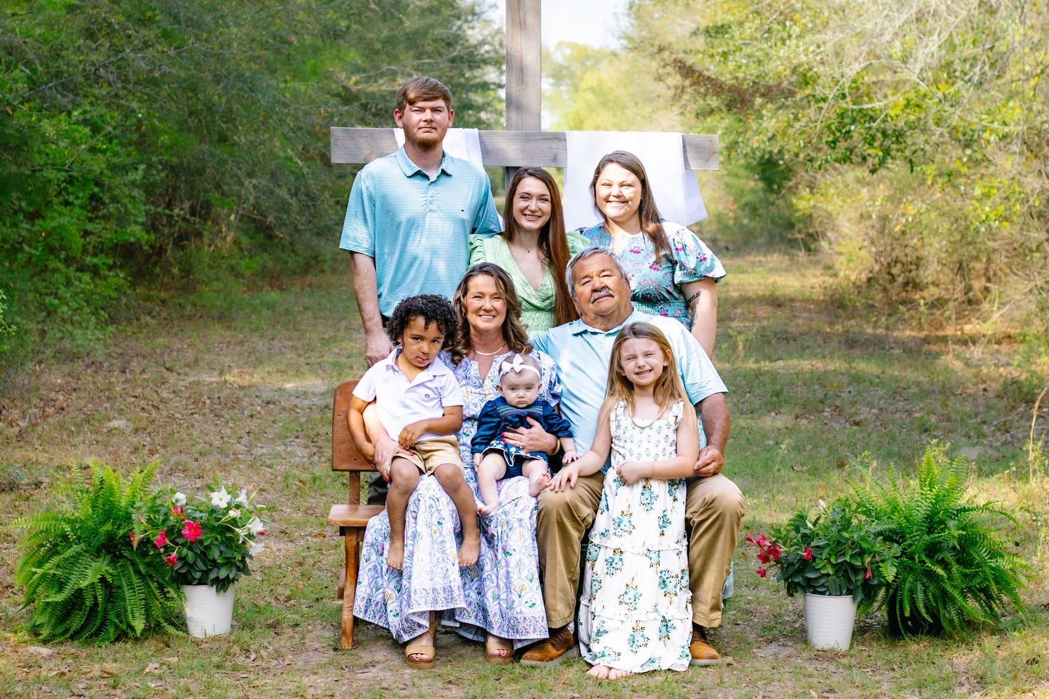 A large family poses together in front of a white cross with ferns outdoors.