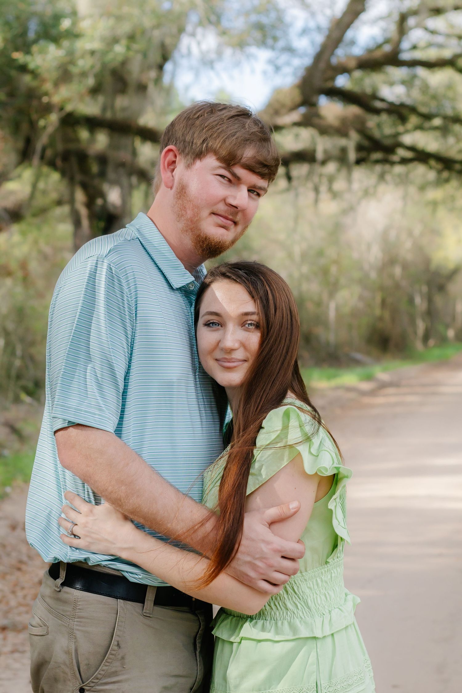 Couple sharing a warm embrace on a tree-lined dirt road, both smiling at the camera.