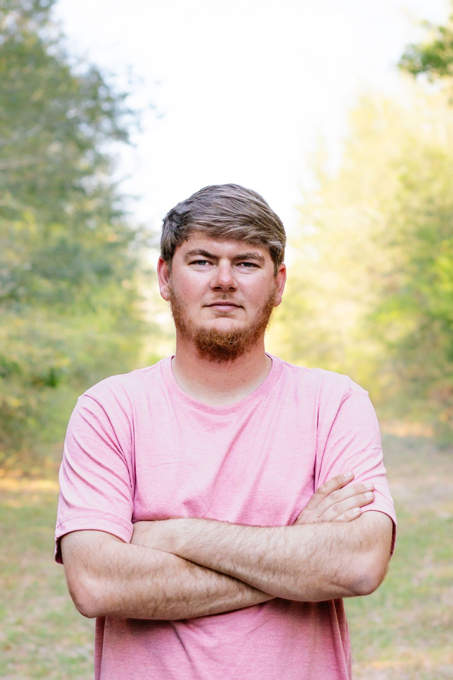 Close-up portrait of a man with beard in a pink shirt standing with arms crossed outdoors.