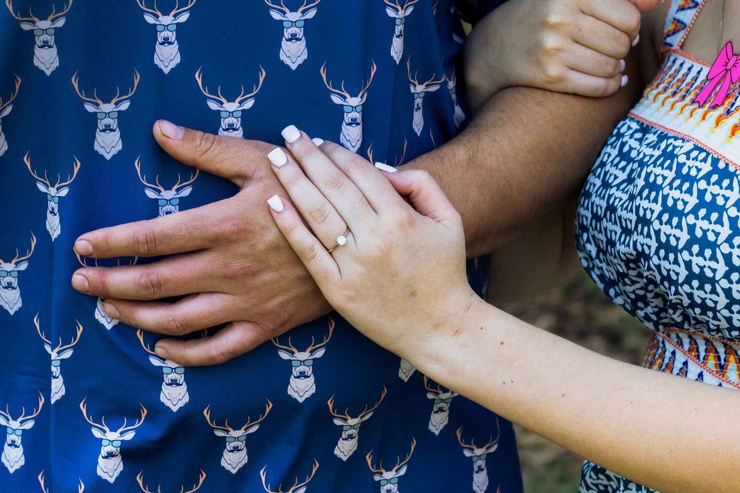 Close-up of couple holding hands showing engagement ring, wearing blue patterned outfits outdoors.