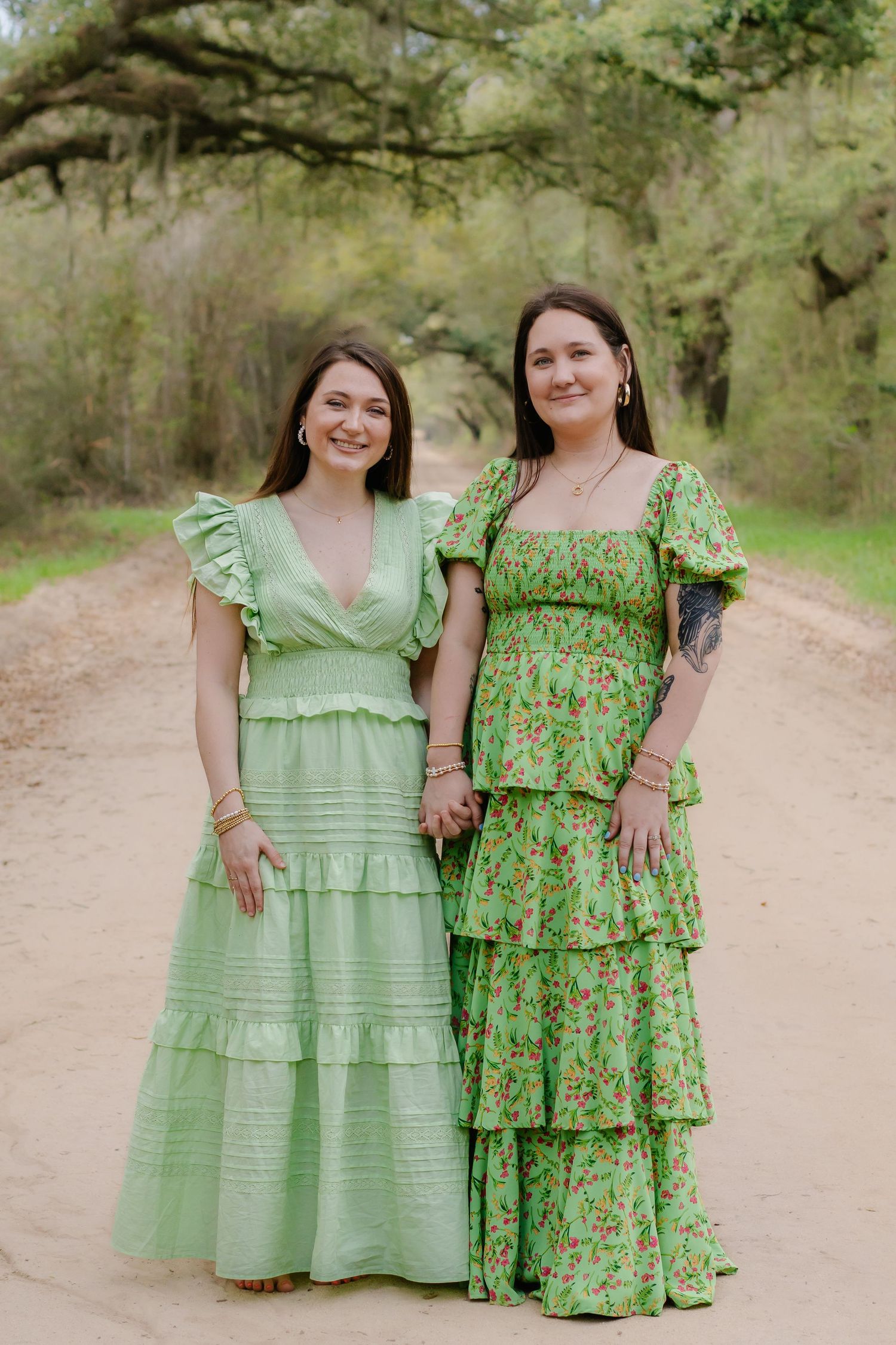 Two young women in green dresses holding hands on a scenic tree-lined dirt road.