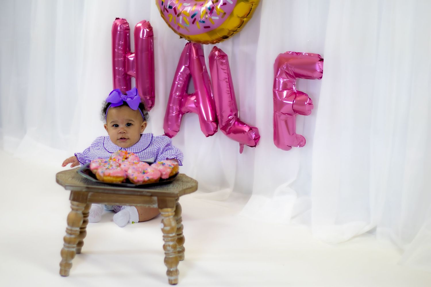 Baby girl in purple bow headband poses with pink frosted donuts on wooden stool at half birthday.