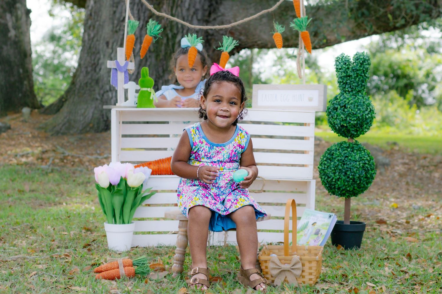 Two girls in floral dresses pose at Easter photo set with topiary and carrot decorations.
