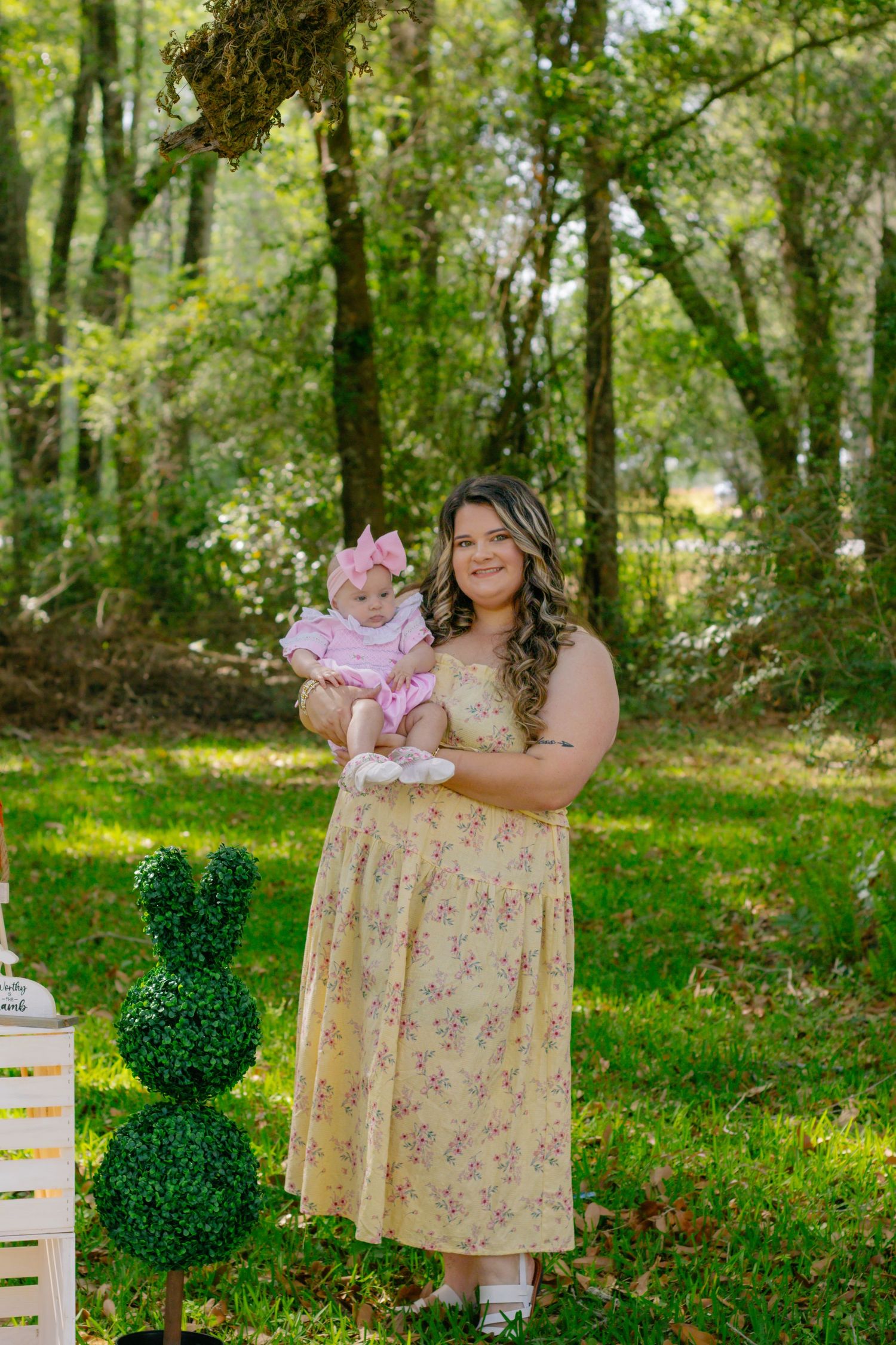 Smiling mother in yellow dress holds baby girl near topiary bunny and white lantern outdoors.