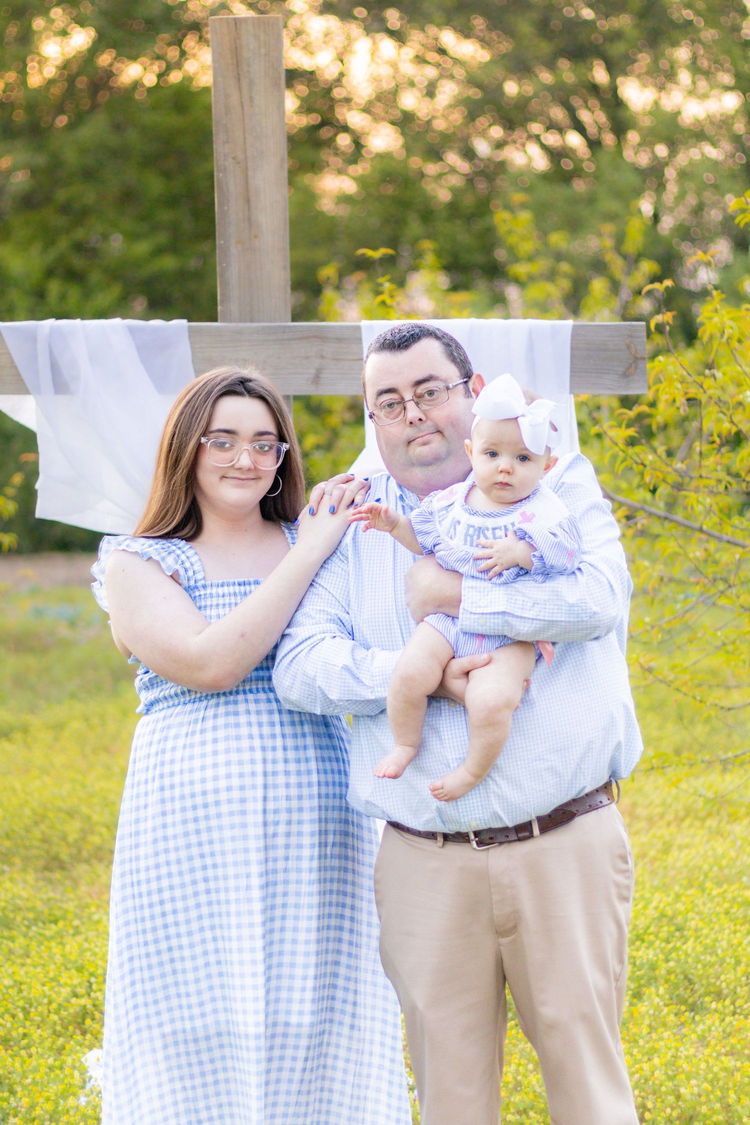 Family of three poses before wooden cross in green field for Easter portrait.