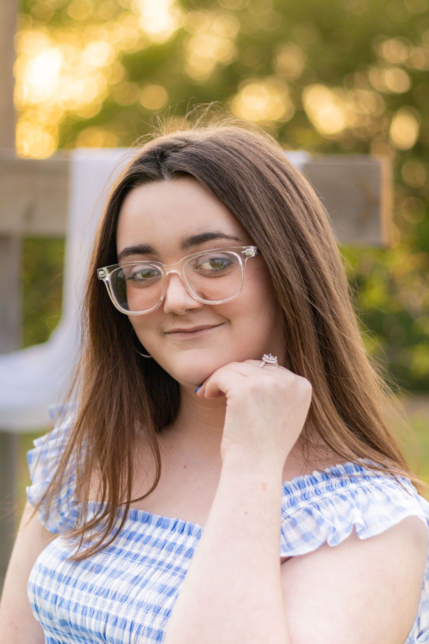 Young woman with long brown hair and clear glasses smiling outdoors in blue gingham off-shoulder top.