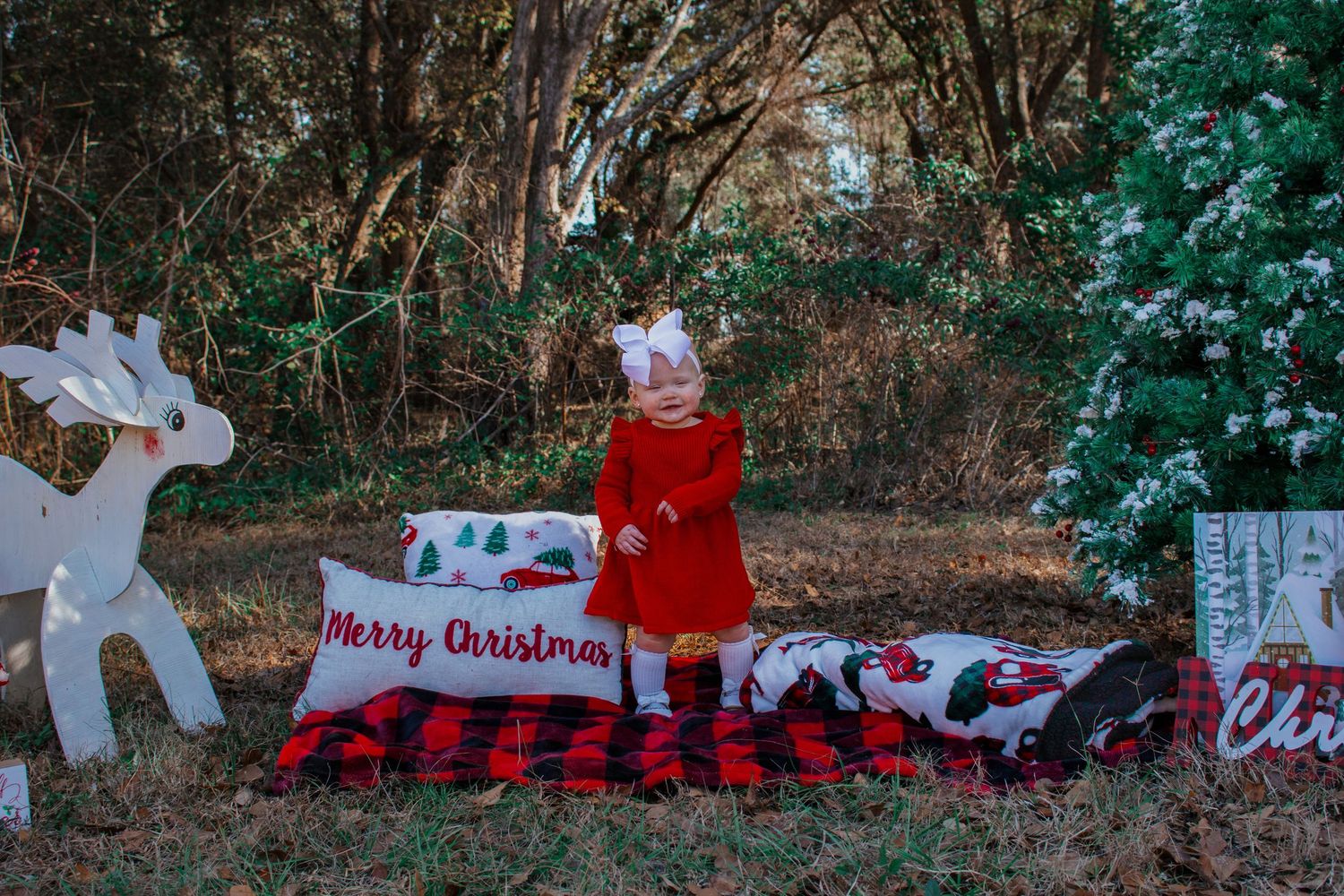 Toddler girl in red dress with bow standing beside Merry Christmas sign in festive outdoor setting.