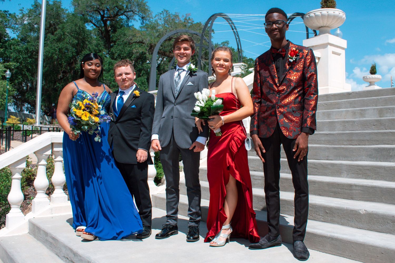Five prom attendees pose on white steps, wearing formal attire including blue and red gowns and suits.