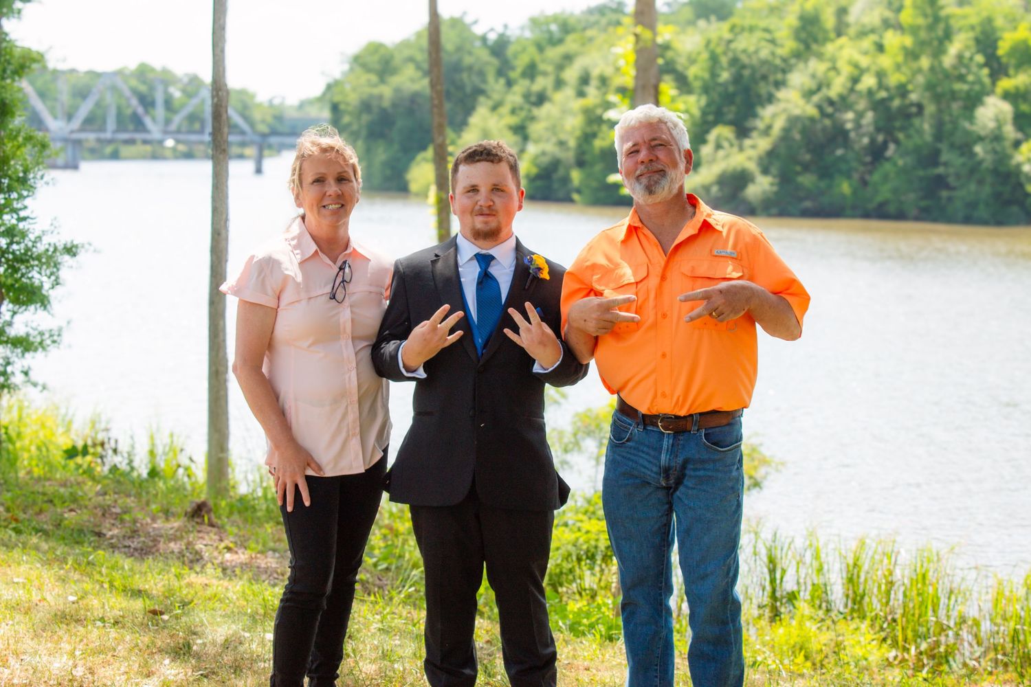Three people pose playfully near a river, with a young man in a suit flanked by two adults.