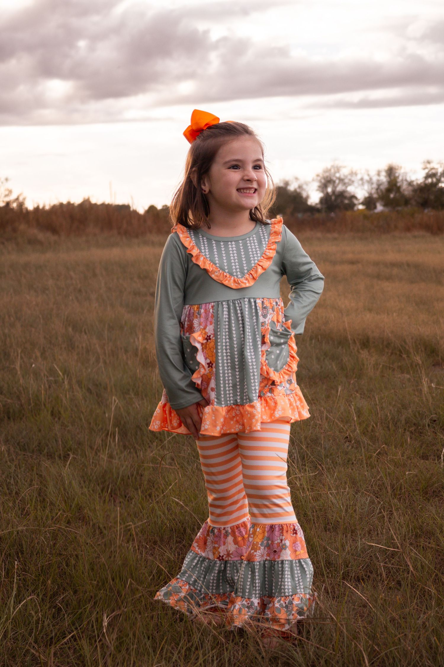 A smiling little girl in orange and teal ruffled outfit with orange bow stands in a field.