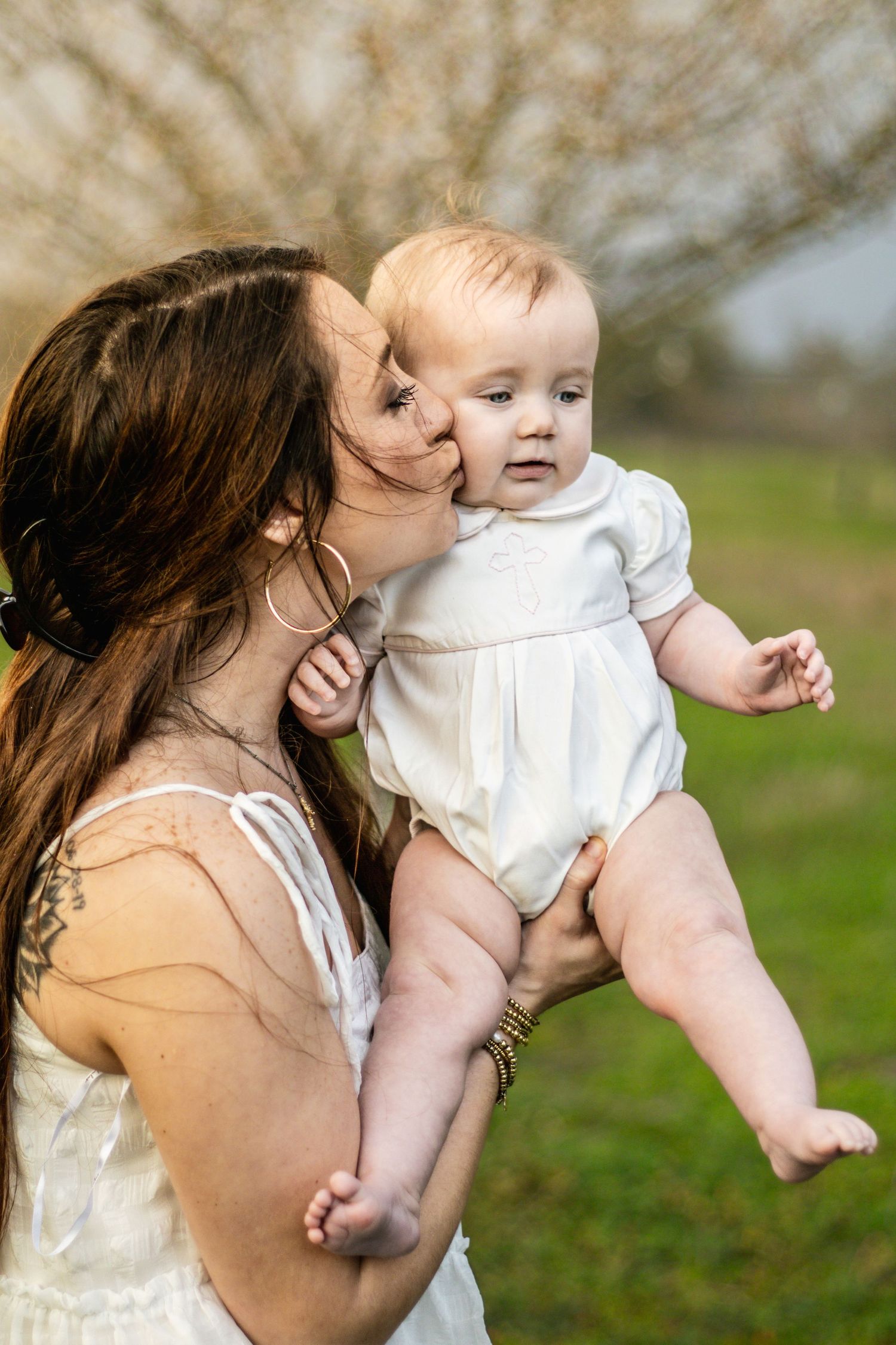 Mother kissing baby in white romper while holding them in a park.