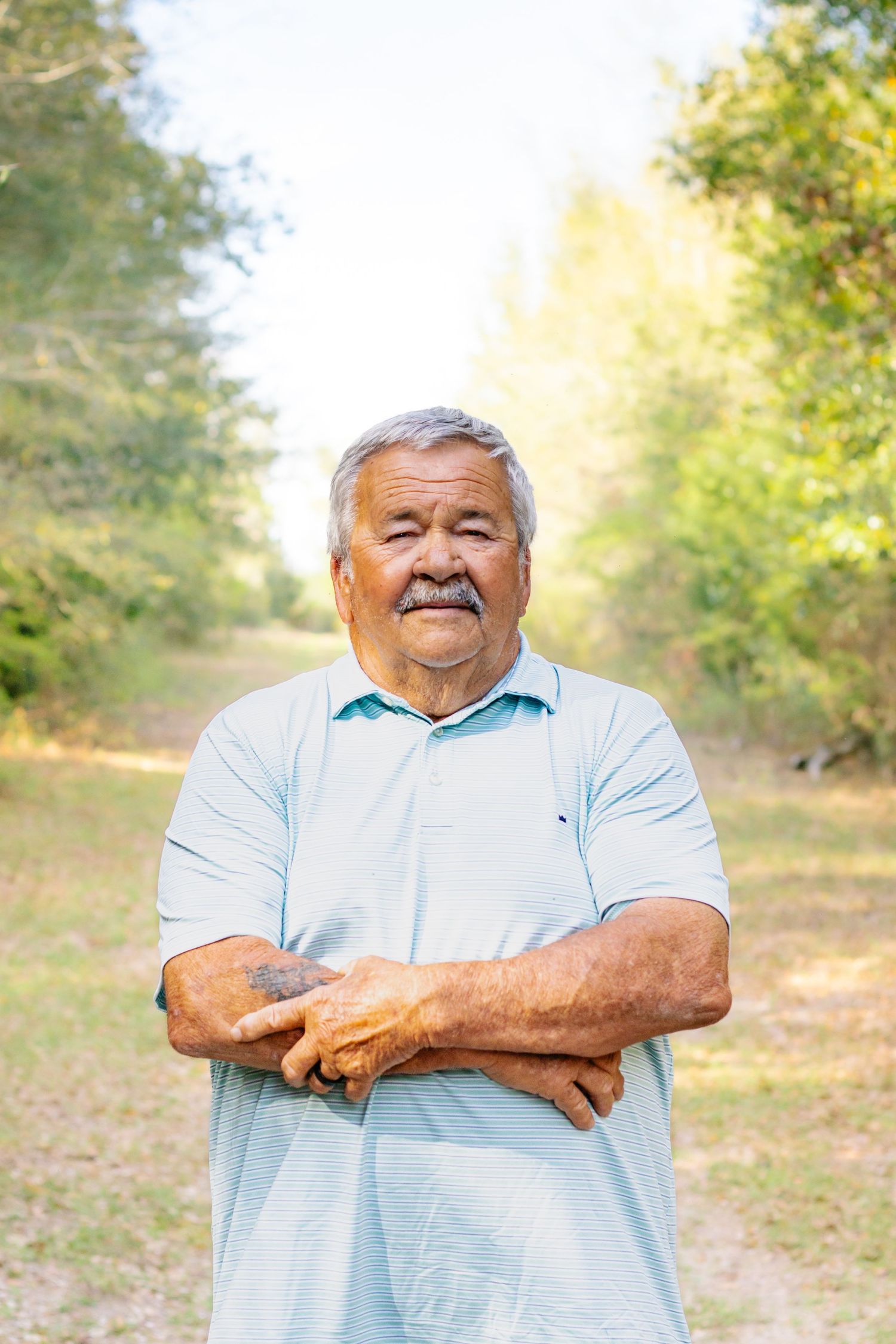 Smiling senior Hispanic man with gray hair and mustache standing outdoors with arms crossed.