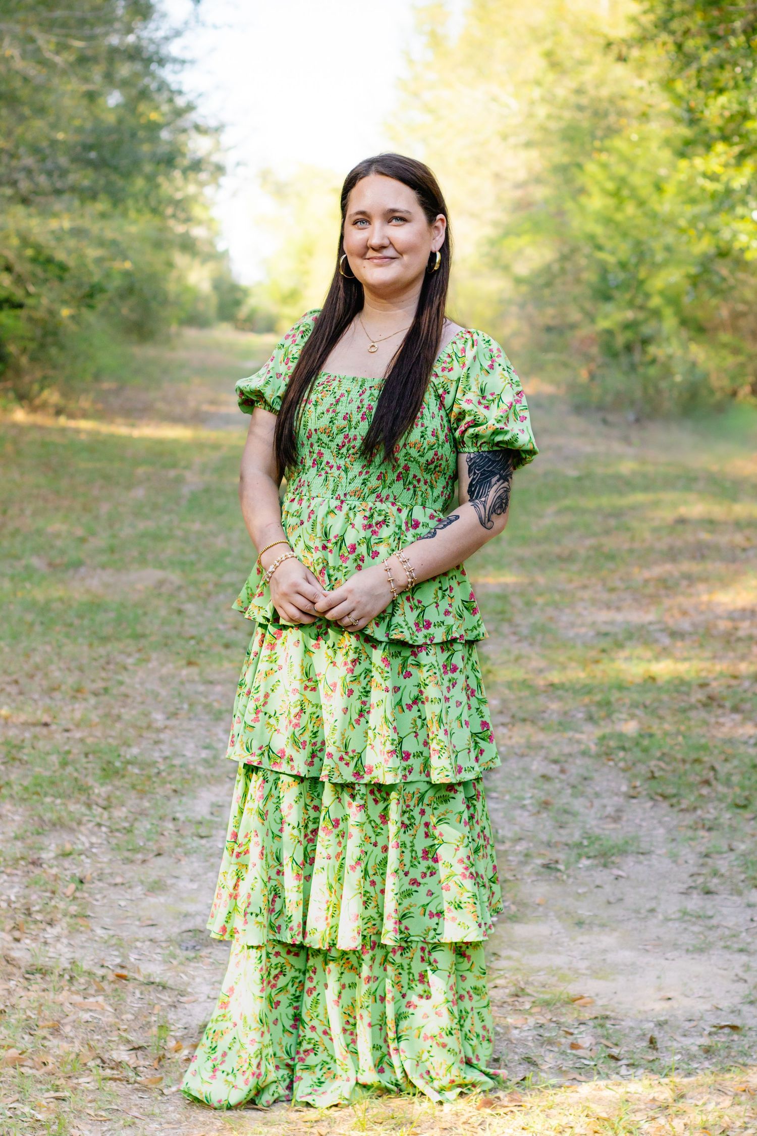 Woman in green tiered floral maxi dress standing on sunlit wooded path outdoors.