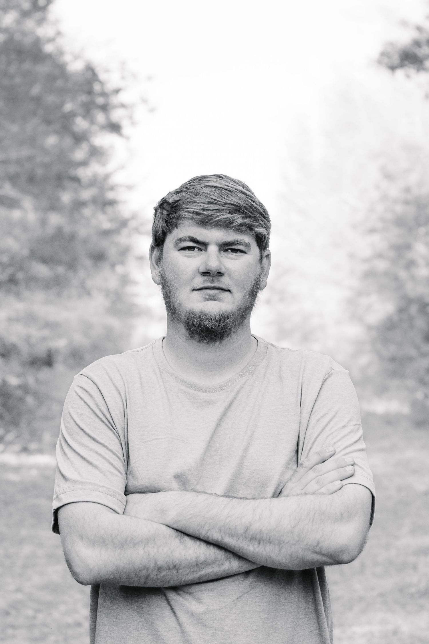 Black and white portrait of a bearded man in a t-shirt standing with arms crossed on a wooded path.
