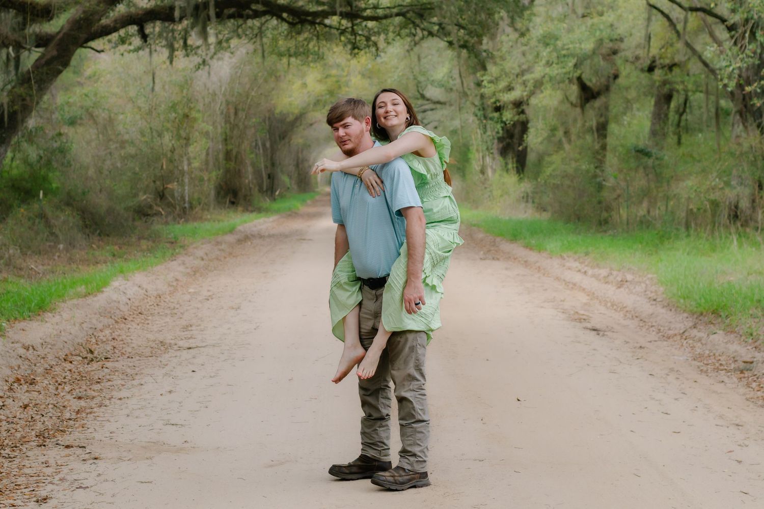 Woman giving man a playful piggyback ride on a tree-lined dirt road during outdoor photo session.