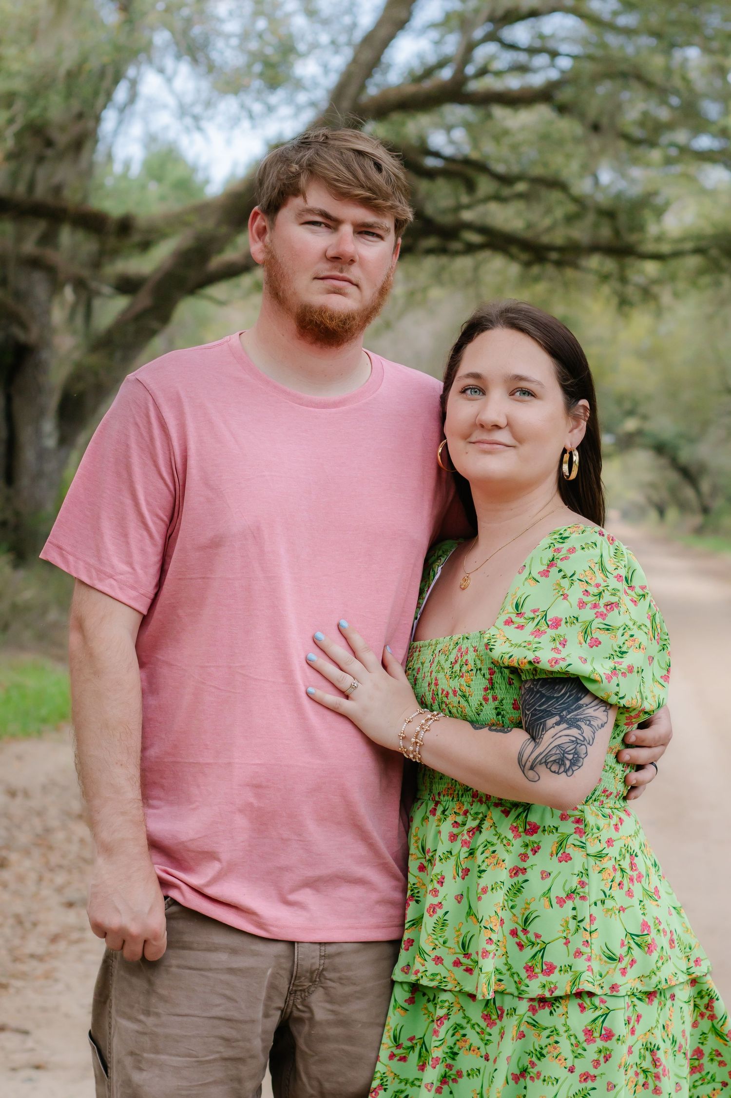 Young couple standing together on a dirt path, man in pink tee and woman in green floral dress embracing.