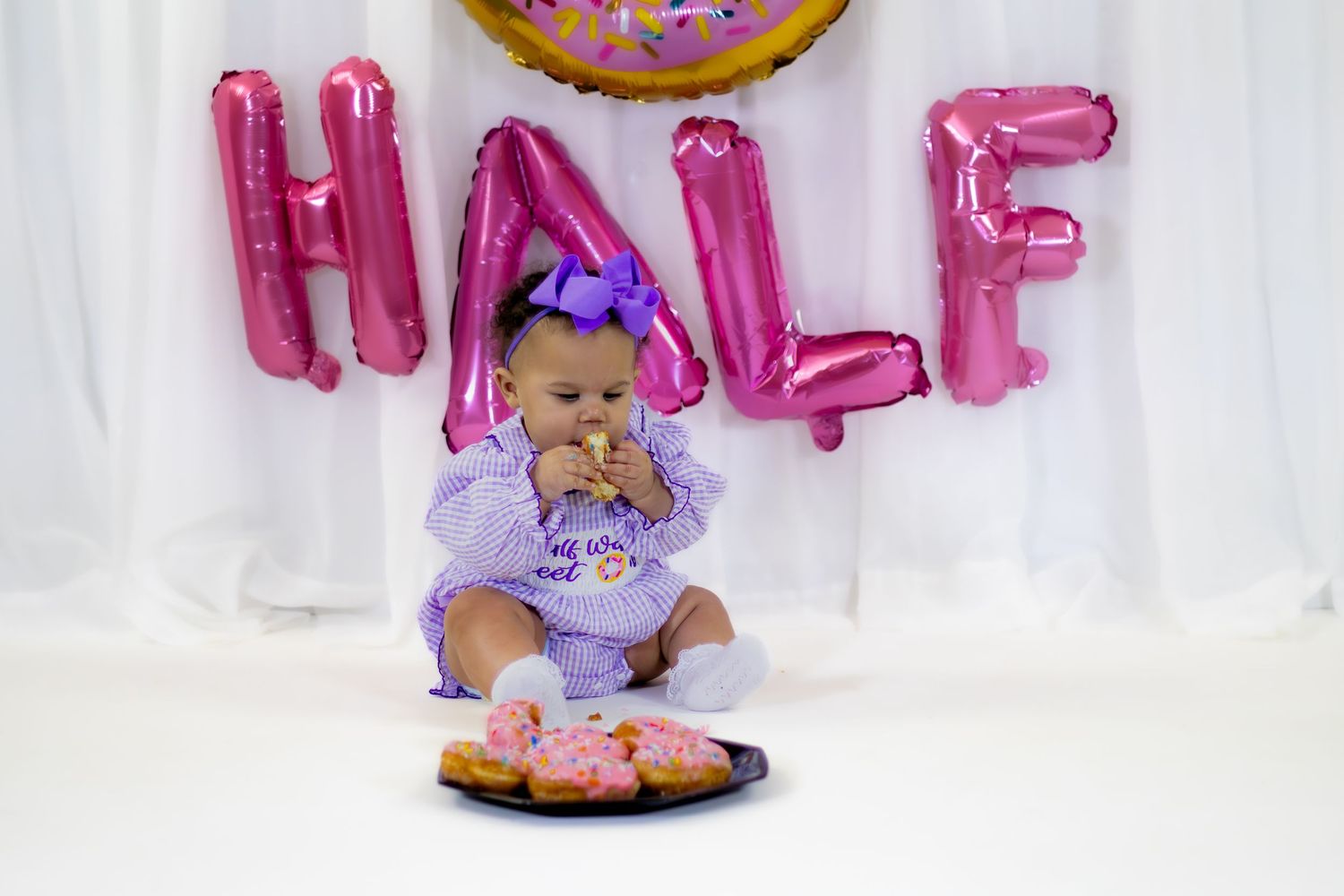 Baby girl in purple outfit reaches for pink frosted donuts at HALF balloon birthday photo shoot.