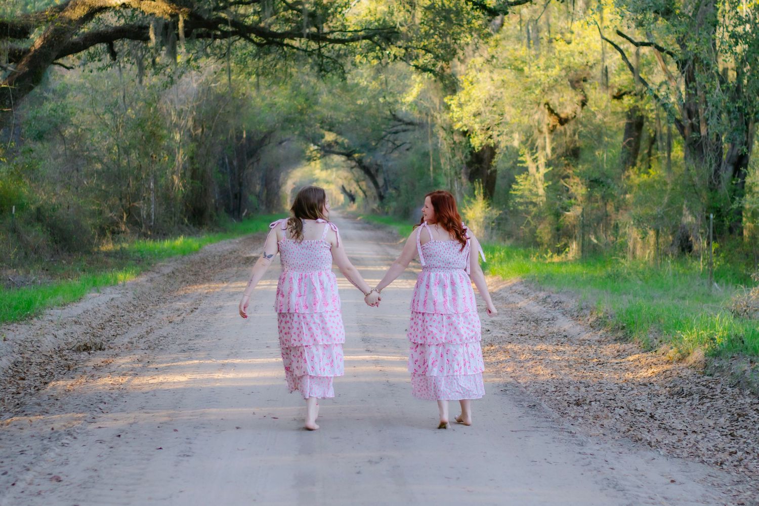 Two girls in pink dresses walking hand-in-hand down a sunlit tree-canopied dirt road.