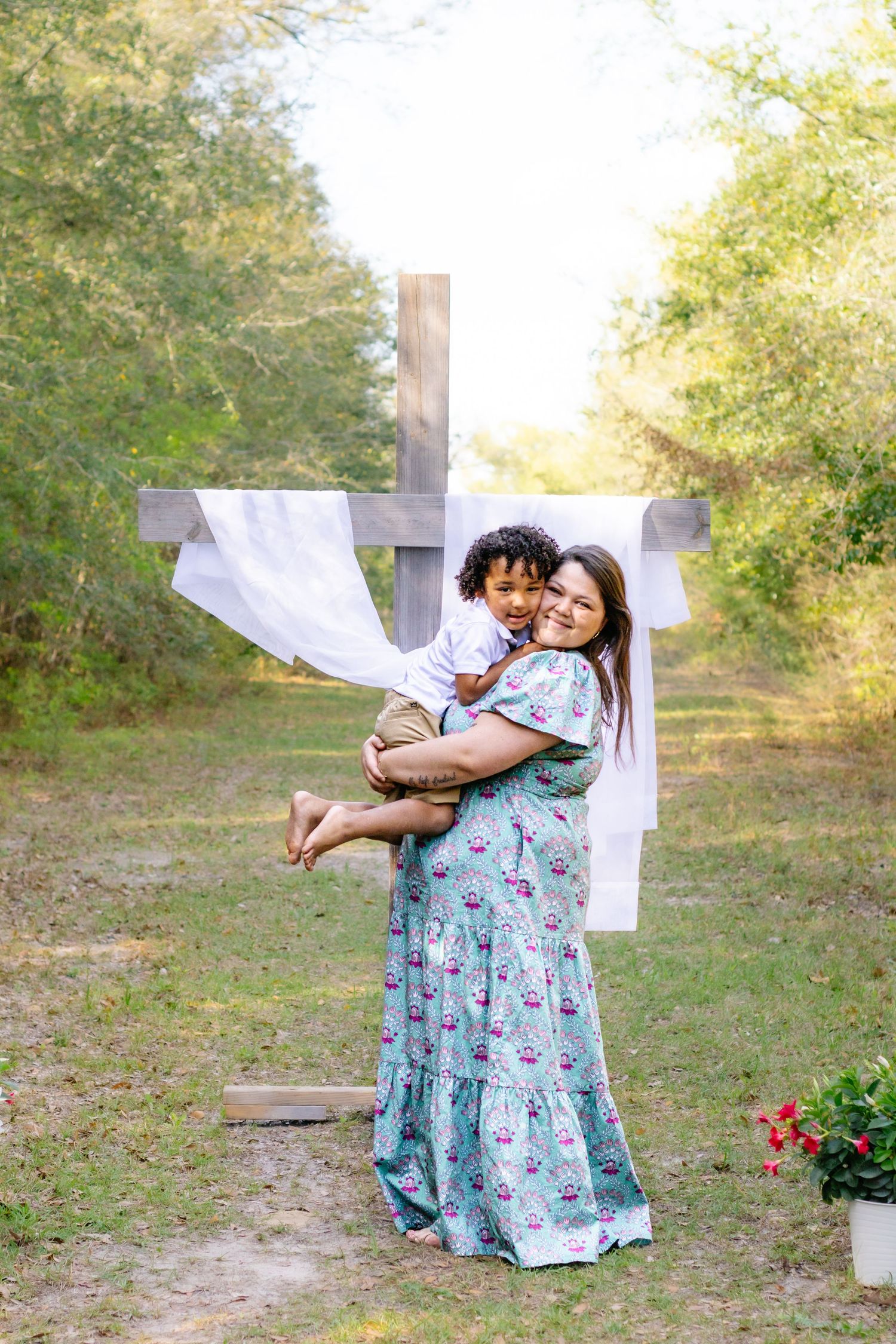 A smiling woman in a floral dress holds a young boy in front of a white-draped wooden cross outdoors.