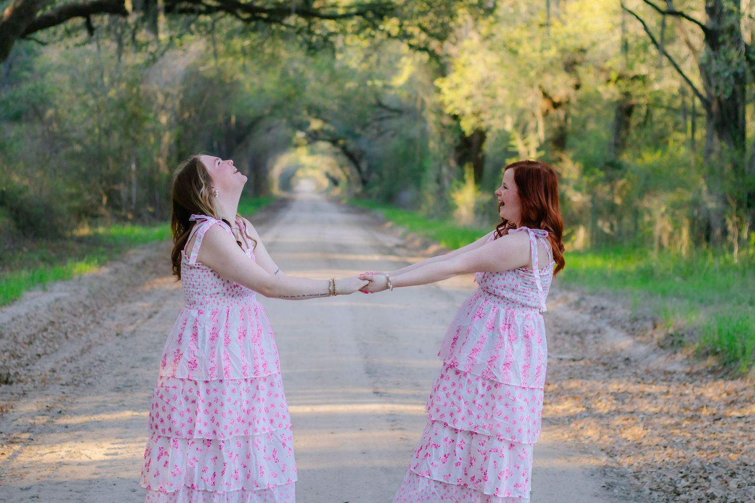 Two girls in matching pink dresses holding hands and spinning on a tree-lined dirt road.
