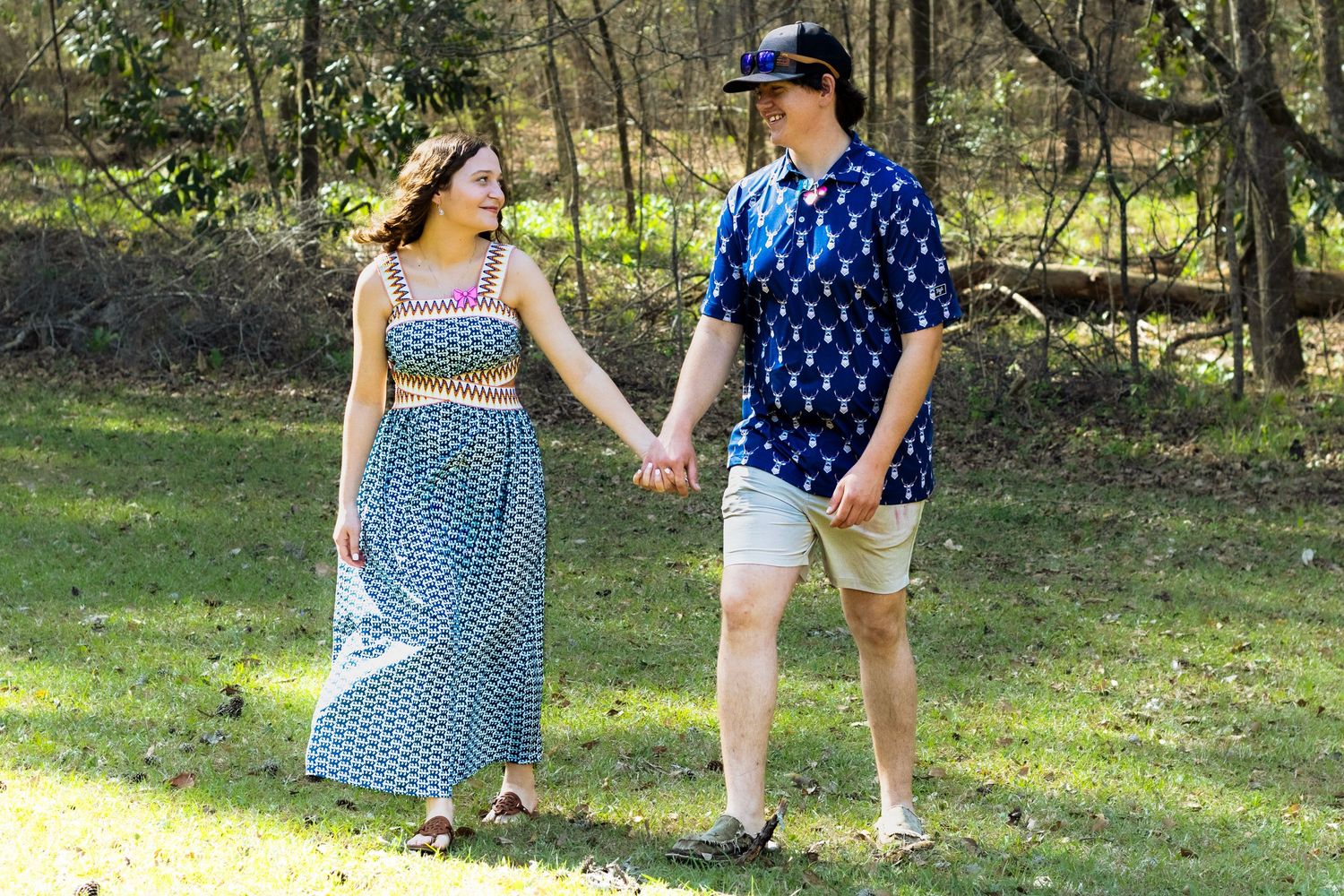 Young couple holding hands walking through sunny green park, woman in blue maxi skirt, man in blue shirt.