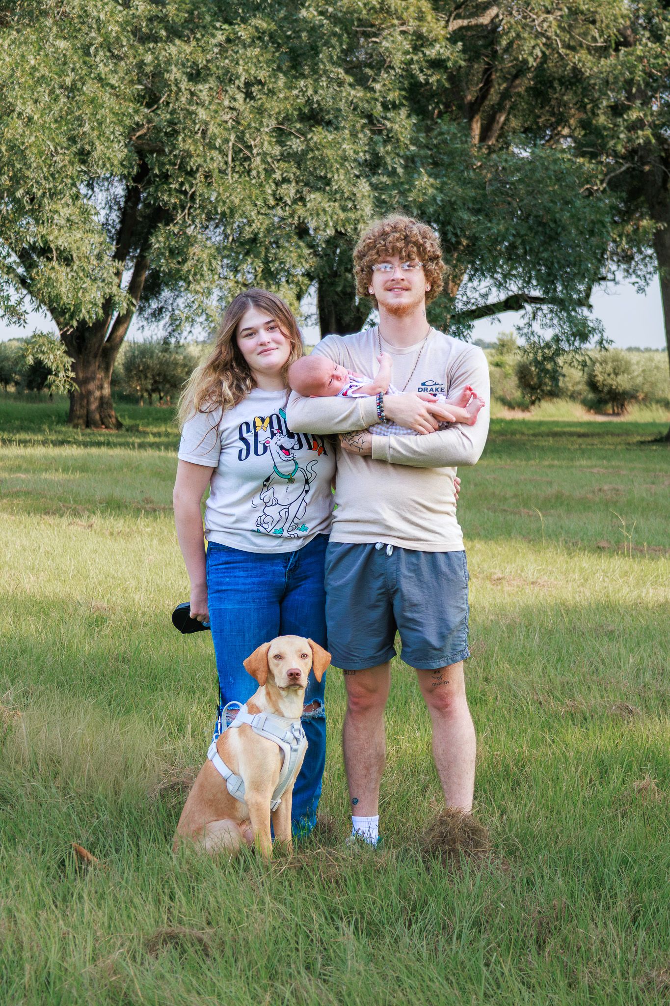 Young couple holding newborn baby with yellow Labrador dog posing outdoors in green field with trees.