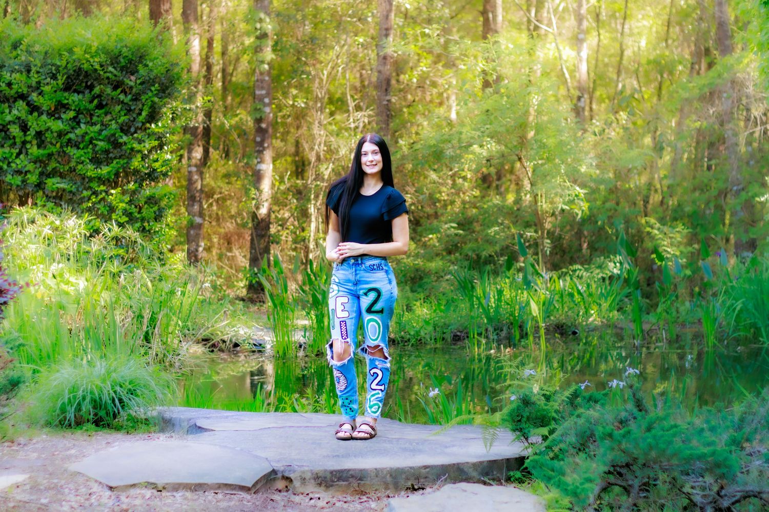 Teen girl in graphic jeans and black top poses on a stone path surrounded by lush green forest and a serene pond.
