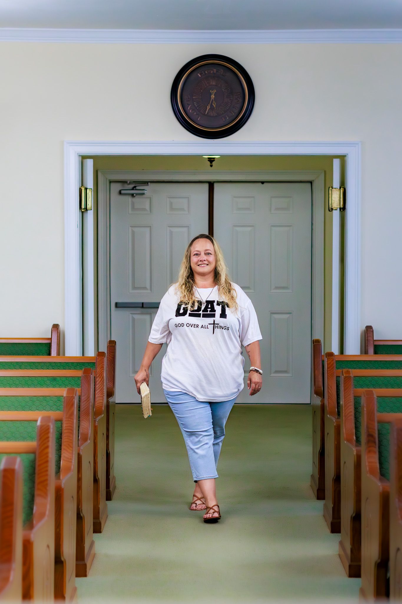 Woman in GOAT shirt walks down church aisle between wooden pews with clock above doorway.
