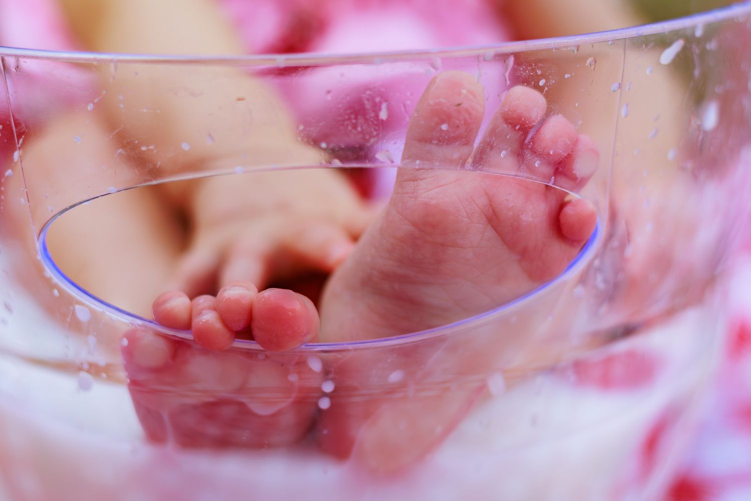 Close-up of newborn baby feet in a clear glass bowl with pink water droplets.
