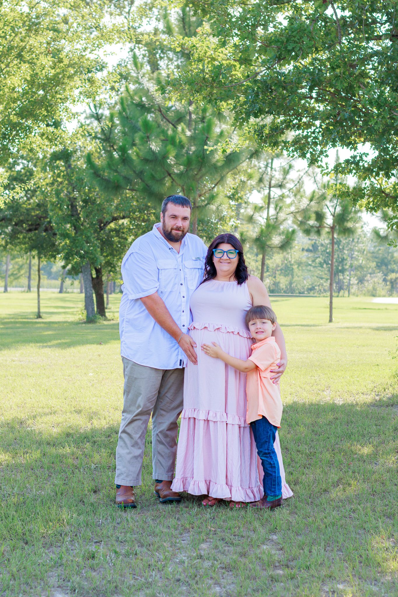 Pregnant woman in pink dress with husband and young son posing outdoors in a lush green park.