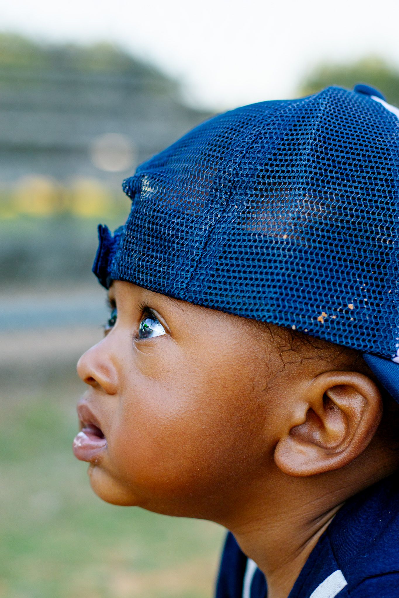 Close-up portrait of a young Black child wearing a blue mesh baseball cap, looking upward with wide eyes.