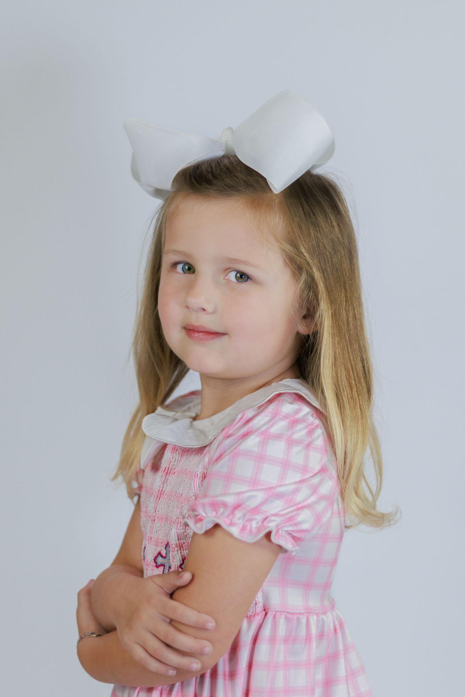 Young girl in pink gingham smocked dress with white bow poses with arms crossed.