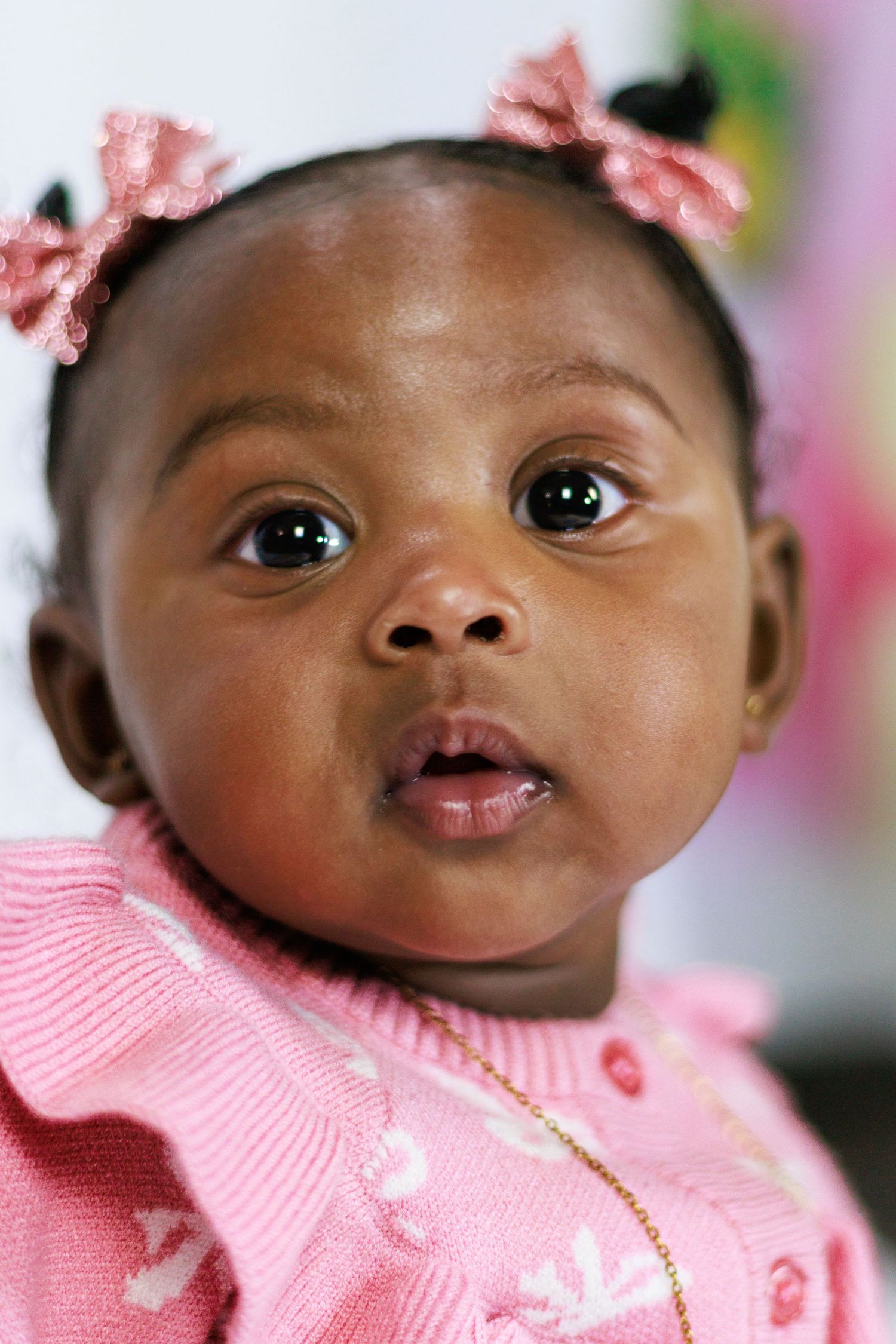 Adorable Black baby girl in pink knit sweater and pink hair bows looks curiously at camera.