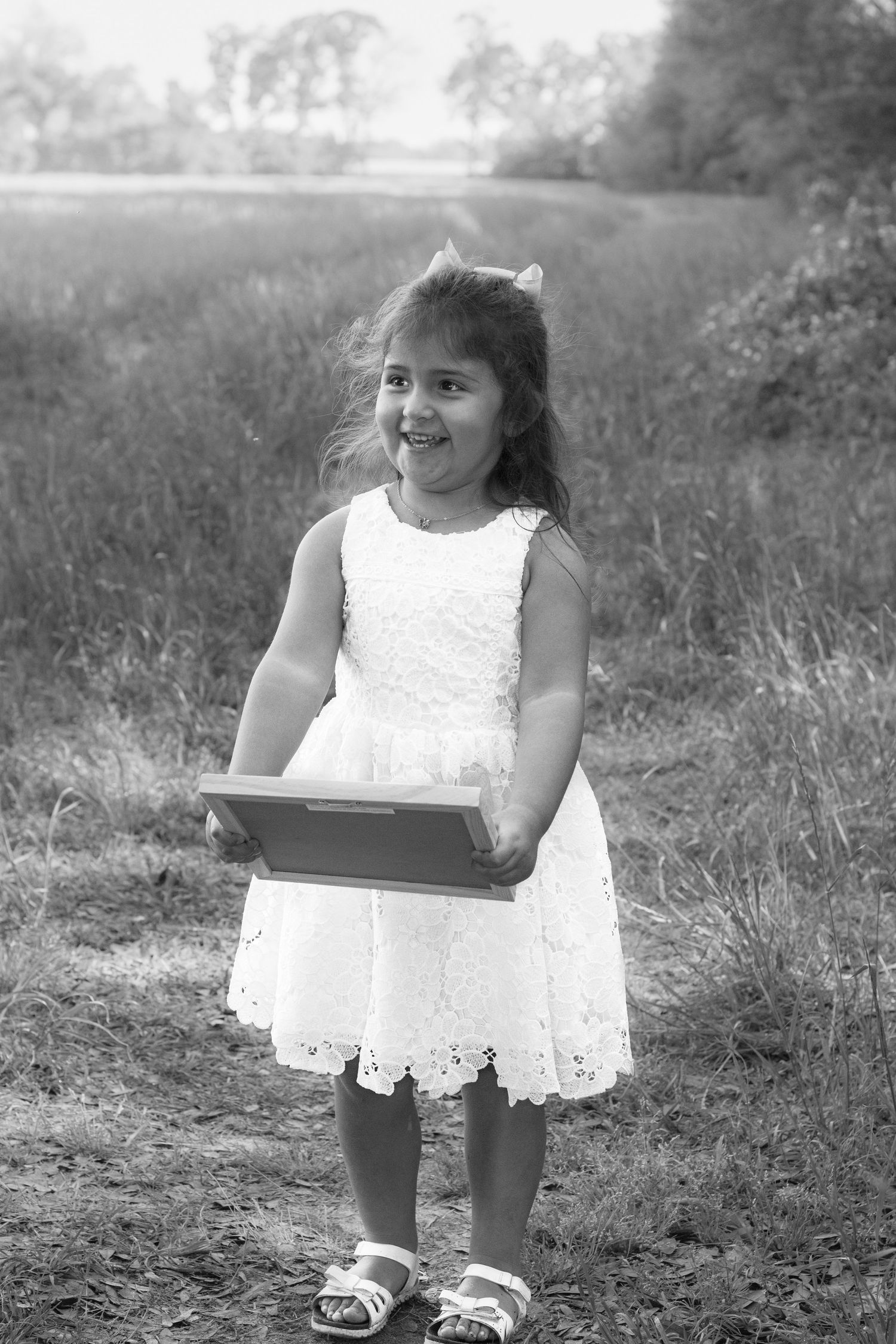 Black and white photo of a smiling little girl in white lace dress holding a tablet outdoors in a field.
