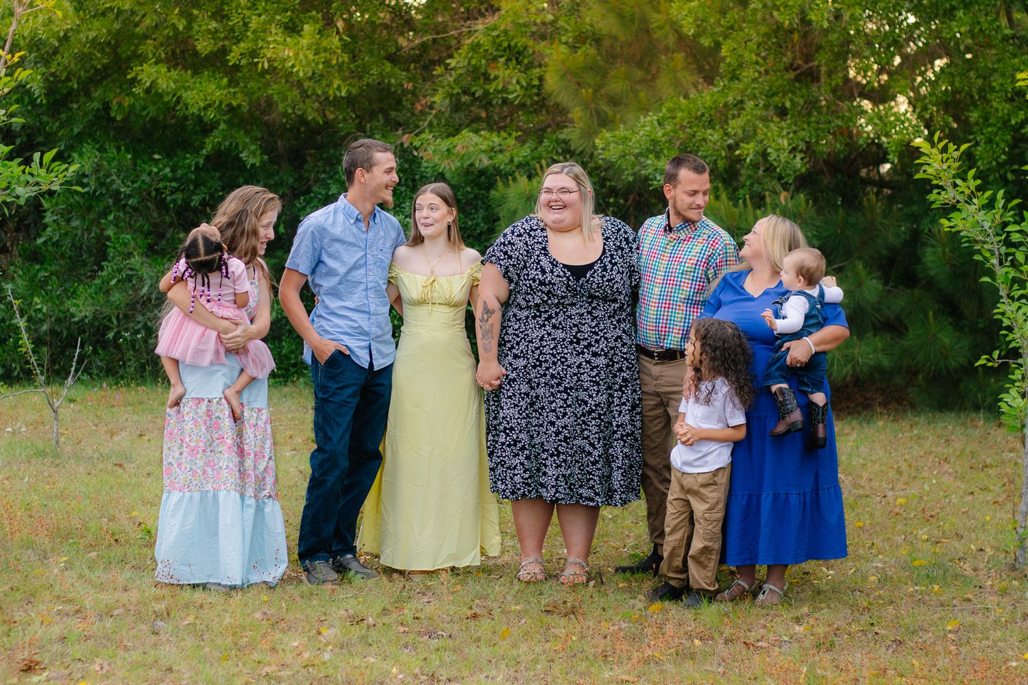 A large family group of eight people poses together outdoors in a lush green backyard setting.