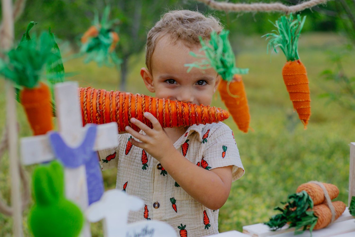 Playful young boy in carrot-print outfit holds a decorative orange carrot near his face at an Easter-themed outdoor setup.