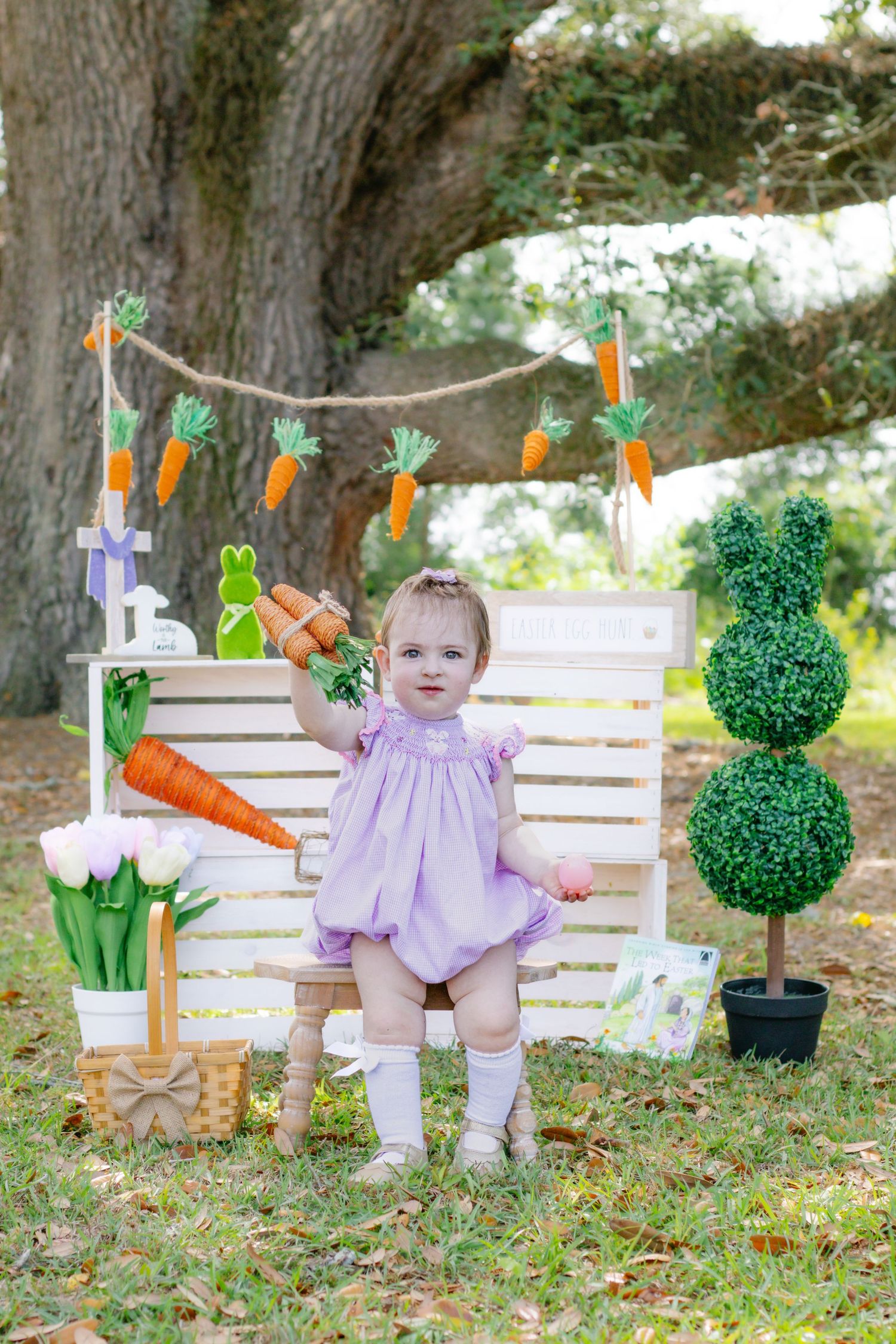 Baby girl in purple romper holding carrot beside Easter decorations with topiary bunny and carrot garland.