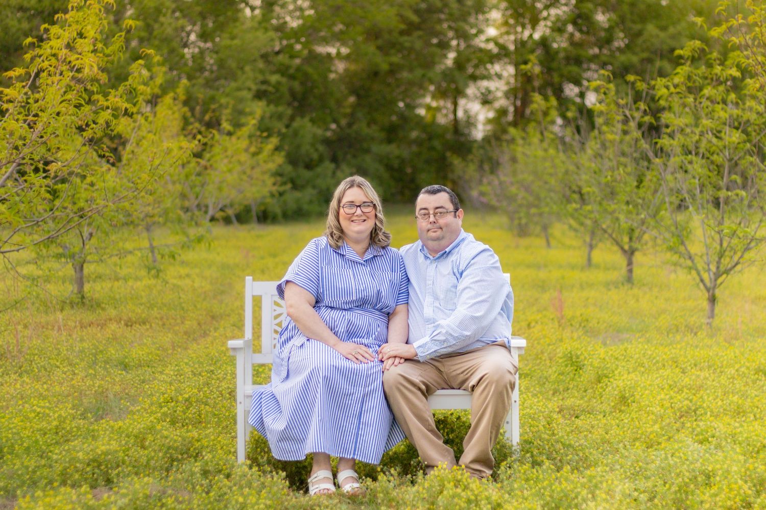 Smiling couple sitting together on white decorative bench surrounded by lush green meadow.