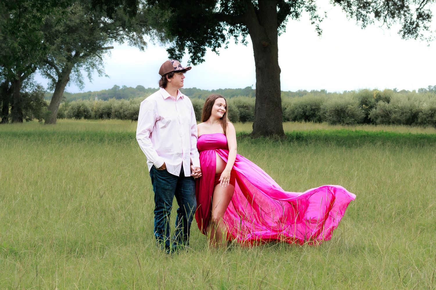 Pregnant woman in flowing pink gown poses with man in white shirt in lush green field with trees.