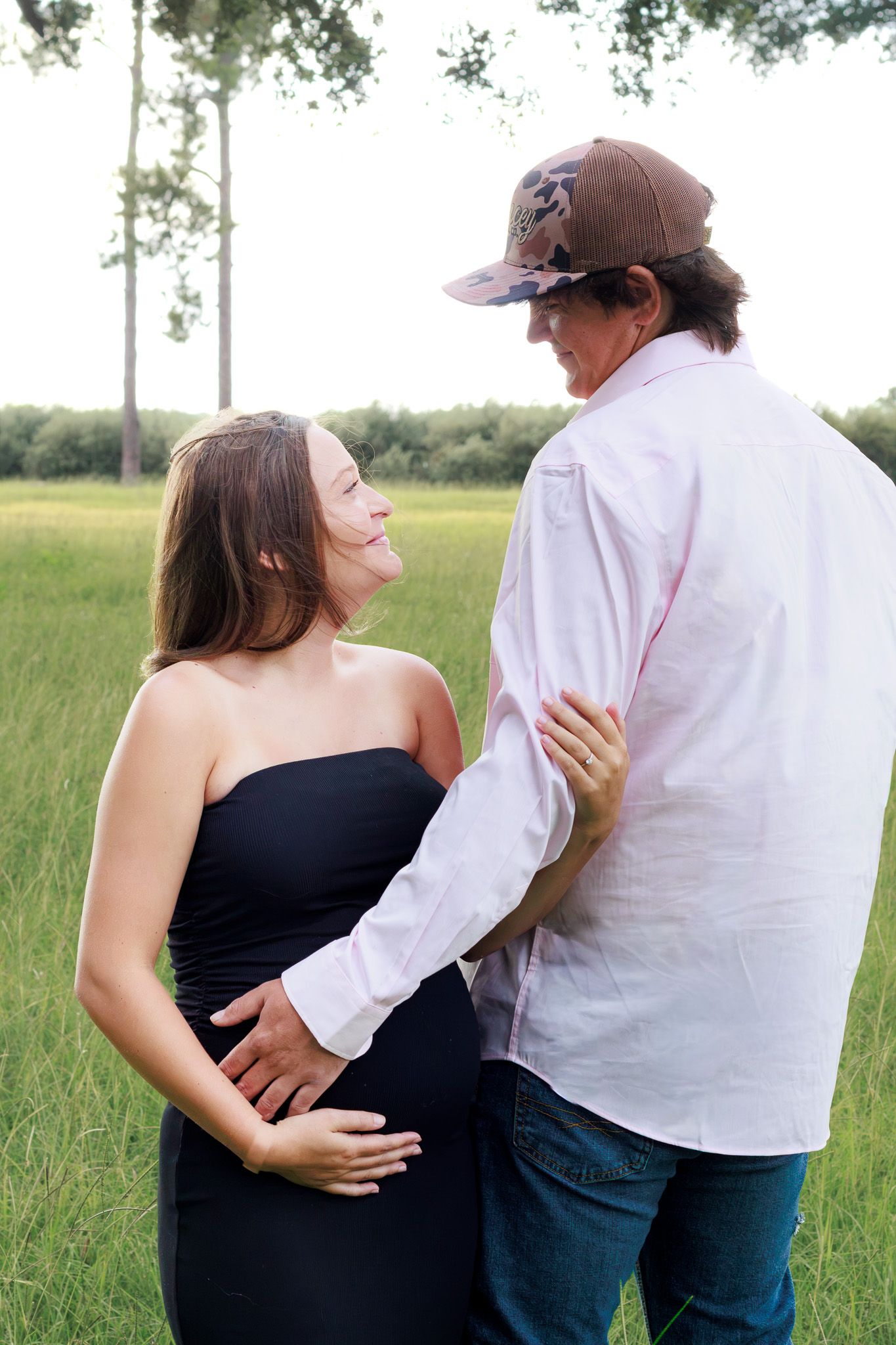 Pregnant woman in black dress with partner in pink shirt and cap standing in green field.