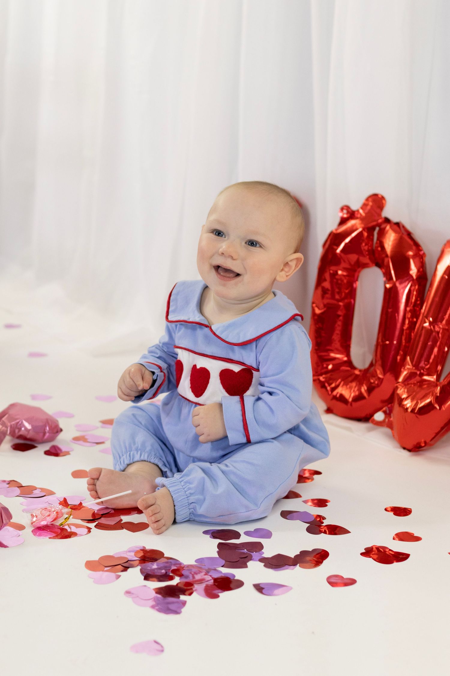 Laughing baby in blue heart romper sits among heart confetti beside red Valentine balloon letters.