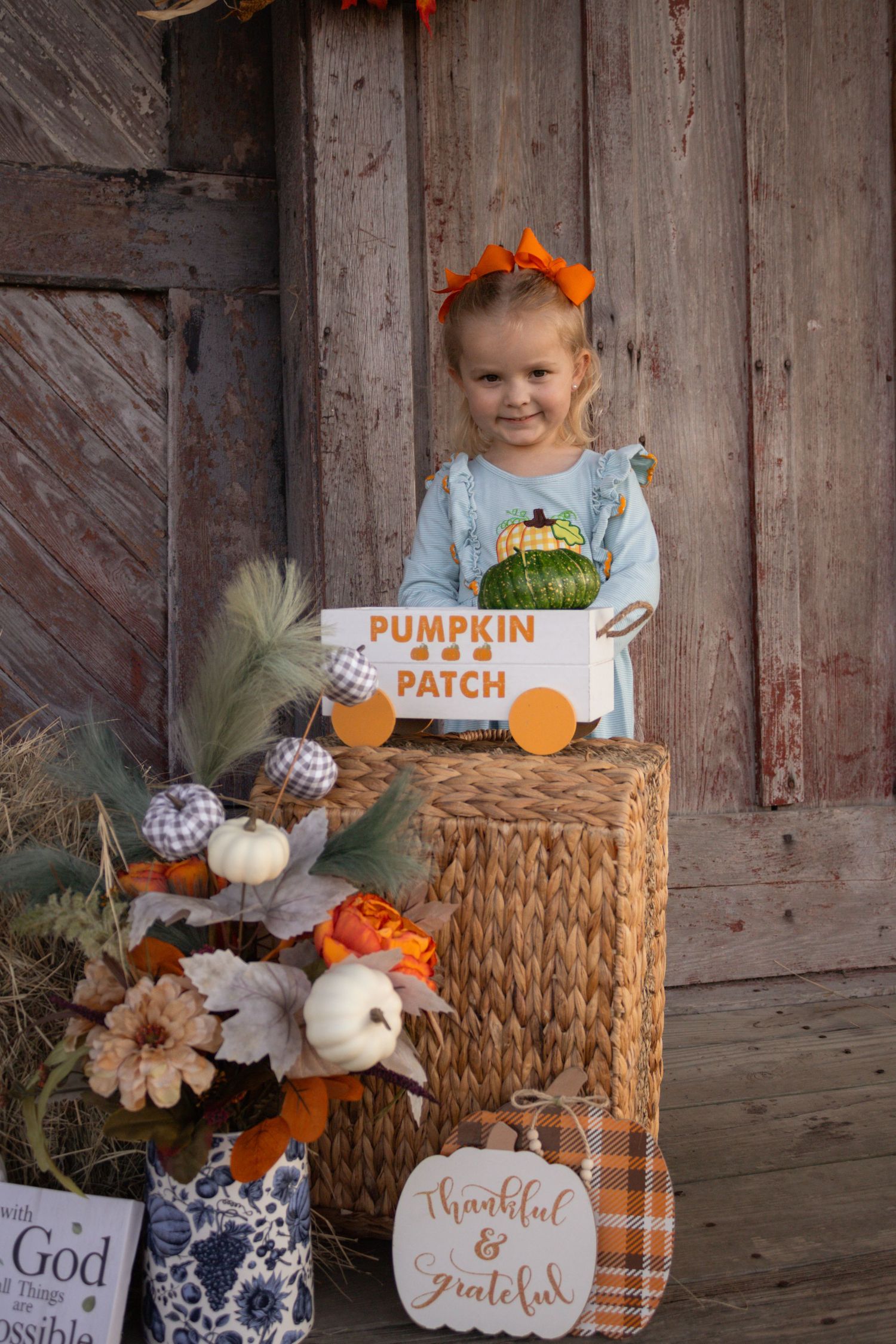 Smiling girl holds a Pumpkin Patch sign while standing behind a wicker basket with autumn decor.