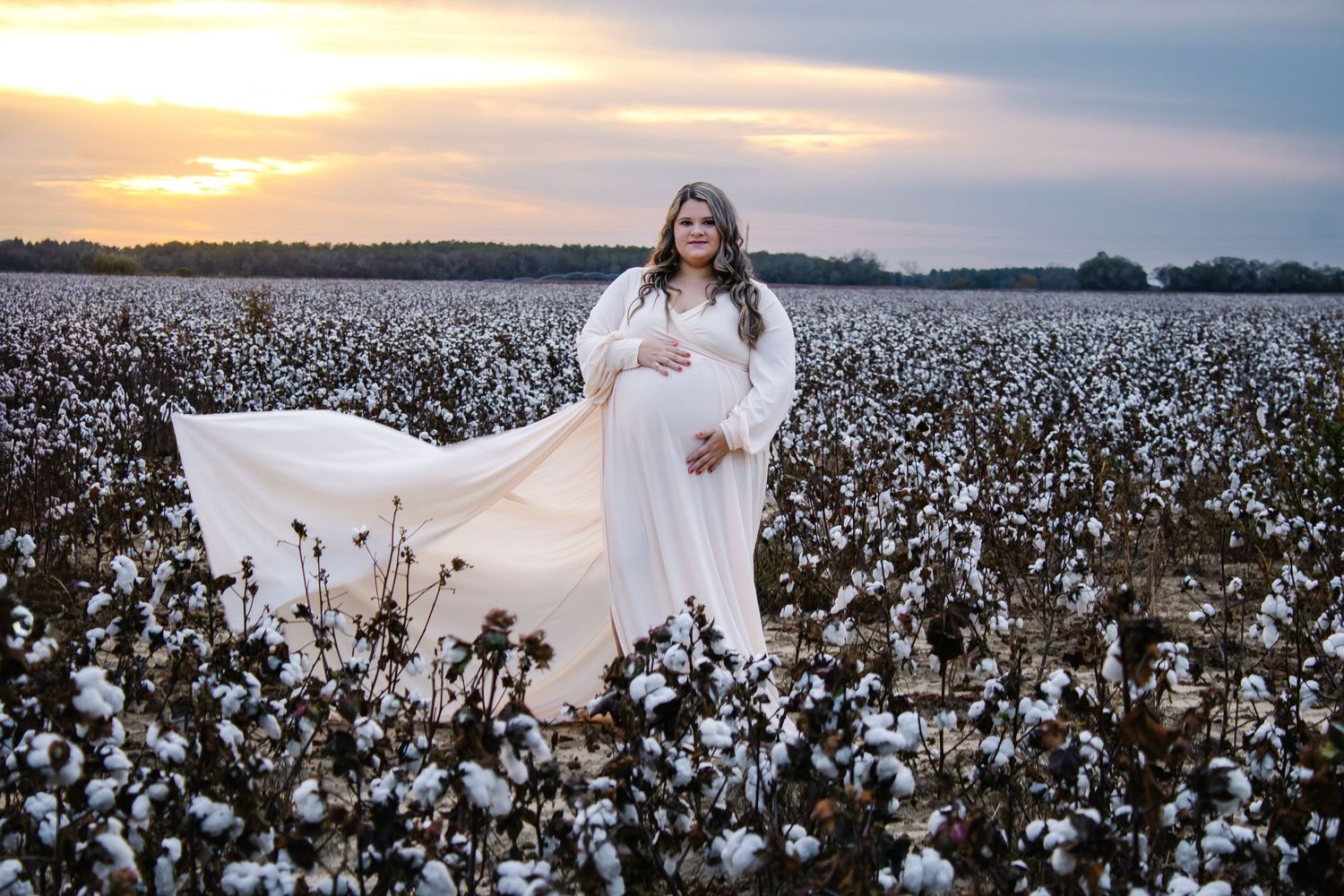 Pregnant woman in white dress with flowing fabric in cotton field at dusk.