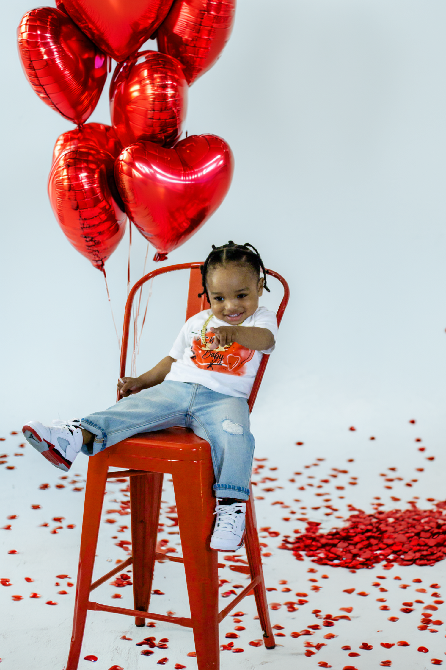 Smiling toddler sits on red chair with red heart balloons and confetti for Valentine's Day portrait.