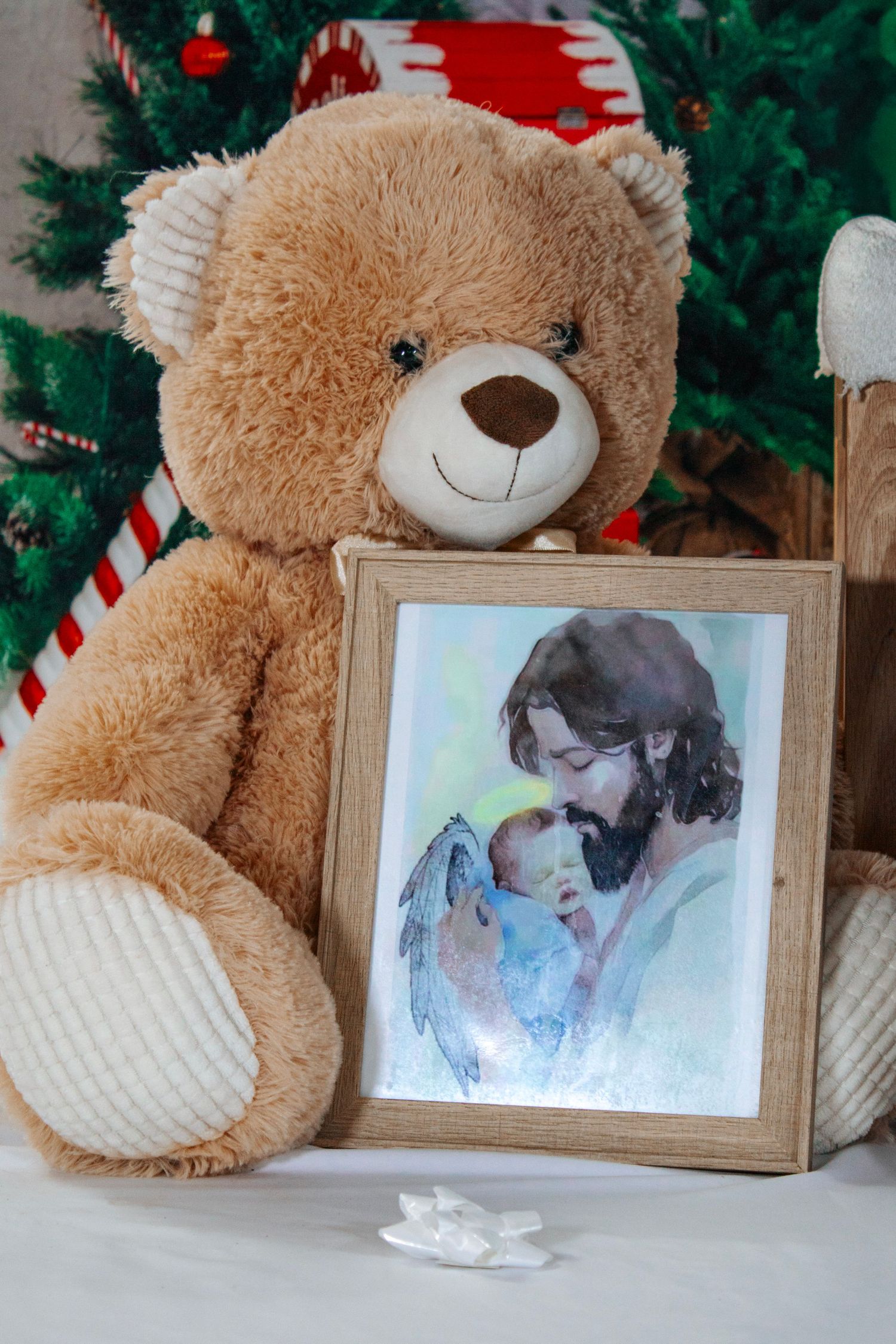 A large teddy bear holds a framed watercolor painting of Jesus with a baby near a Christmas tree.